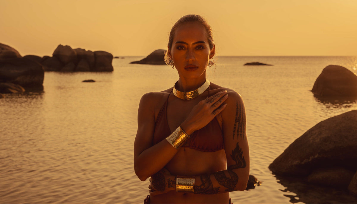 A woman stands in shallow water at sunset wearing a dark red bikini top and bottom, accessorized with a white collar necklace, gold cuff bracelet, and a gold belt. Large rocks are visible in the water around her.
